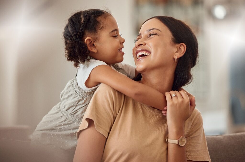 Eine glückliche Mutter mit ihrer kleinen Tochter, eine Umarmung zu Hause und ein Lebensstil mit Spielzeit am Wochenende für die Mutter als Elternteil. Ein lustiges, ausgelassenes Kind im Wohnzimmer, die Mutter hält ihr Kleinkind mit einem Lächeln im Arm, und am Muttertag wächst die Bindung zwischen ihnen